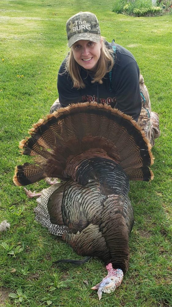 Executive Assistant, Liz Shelley, poses with a big smile on bright green grass, holding a large turkey she harvested.